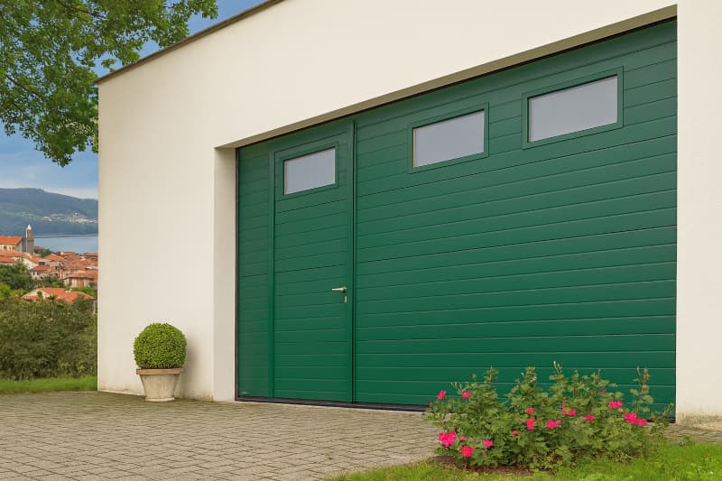 Puerta seccional verde con puerta peatonal integrada y ventanas rectangulares, instalada en garaje moderno de San Vicente do Mar (Pontevedra)